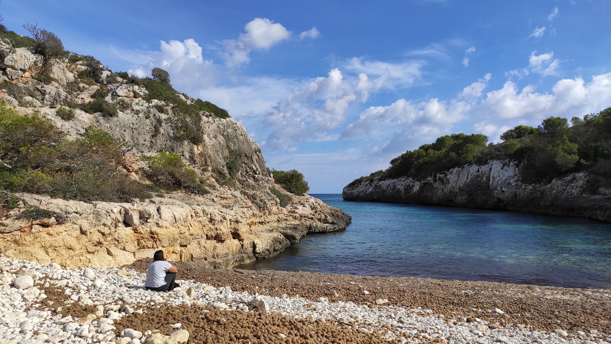 Person sitzt auf steinigem Strand und blickt in türkisfarbene Bucht zwischen bewachsenen Klippen unter blauem Himmel.