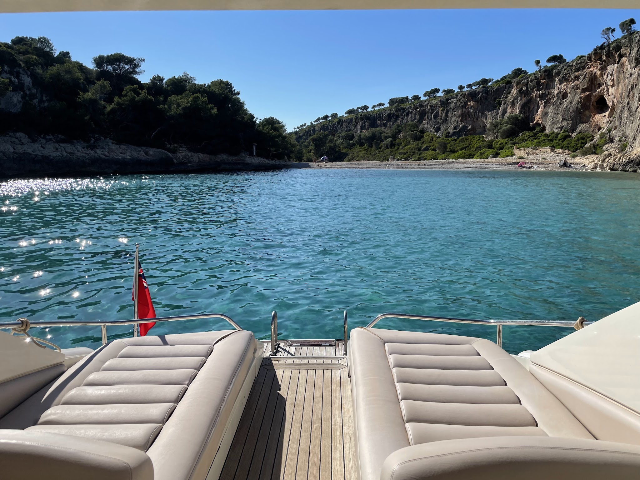 Zwei gepolsterte Liegen auf Yachtheck blicken auf funkelnd türkisene Bucht mit bewaldeten Klippen im Sonnenlicht.