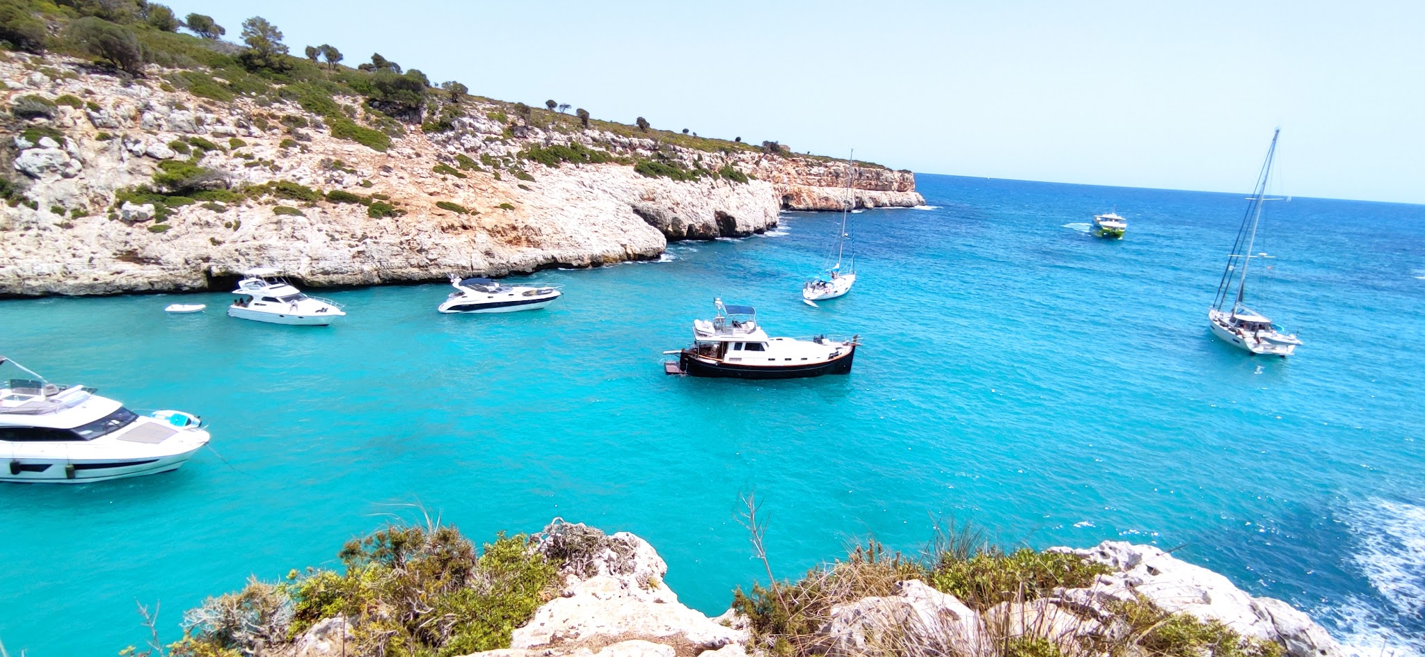 Türkisblaue Bucht mit mehreren vor Anker liegenden Motor- und Segelbooten, umgeben von felsigen, bewachsenen Klippen