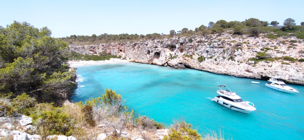 Türkisblaue Bucht mit kleinem Sandstrand, von Felsküste und Pinien umrahmt; zwei weiße Yachten ankern im Wasser.