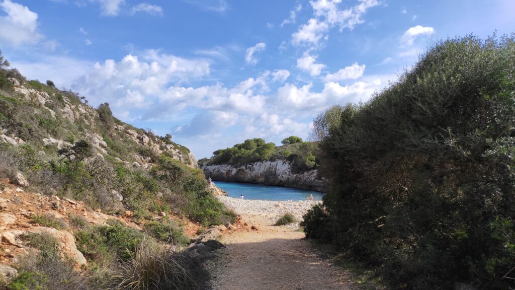 Schotterpfad zwischen buschigen Felsen endet an kleiner Bucht mit türkisblauem Wasser, Badende, teils bewölkter Himmel