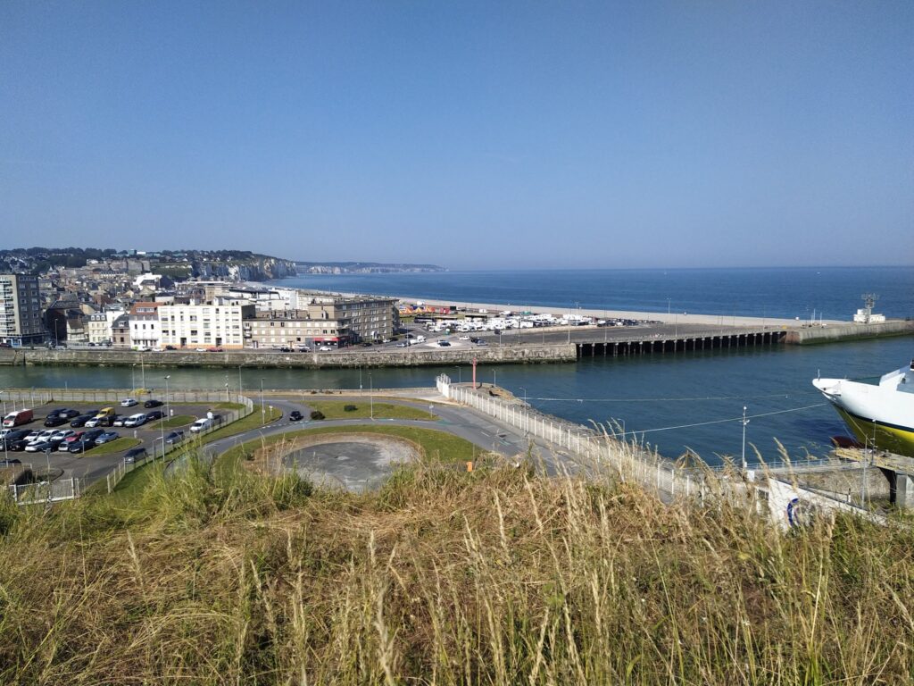 Küstenstadt mit Hafenbecken, parkenden Autos und Fähre; Steilküste und weites Meer unter klarem Himmel.