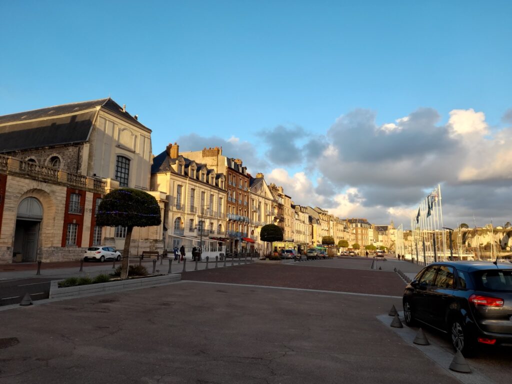Abendsonne erleuchtet historische Häuser am Hafenplatz; Autos, Flanierende und wehende Fahnen im Wind.