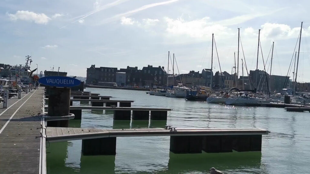 Stiller Hafen: leere Stege mit Schild „Vauquelin“, dahinter Segelboote und Uferhäuser unter leicht bewölktem Himmel.
