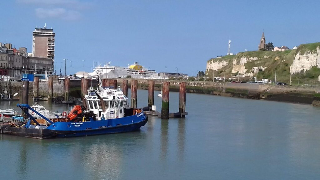Blaues Hafenboot vor Pfählen; dahinter Fähre, Stadtgebäude und weiße Klippe mit Kirche unter blauem Himmel.