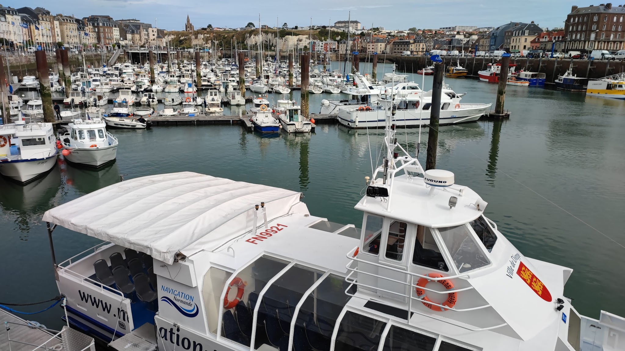 Marina mit vielen weißen Booten; im Vordergrund ein überdachter Ausflugs-Katamaran, Stadthäuser dahinter