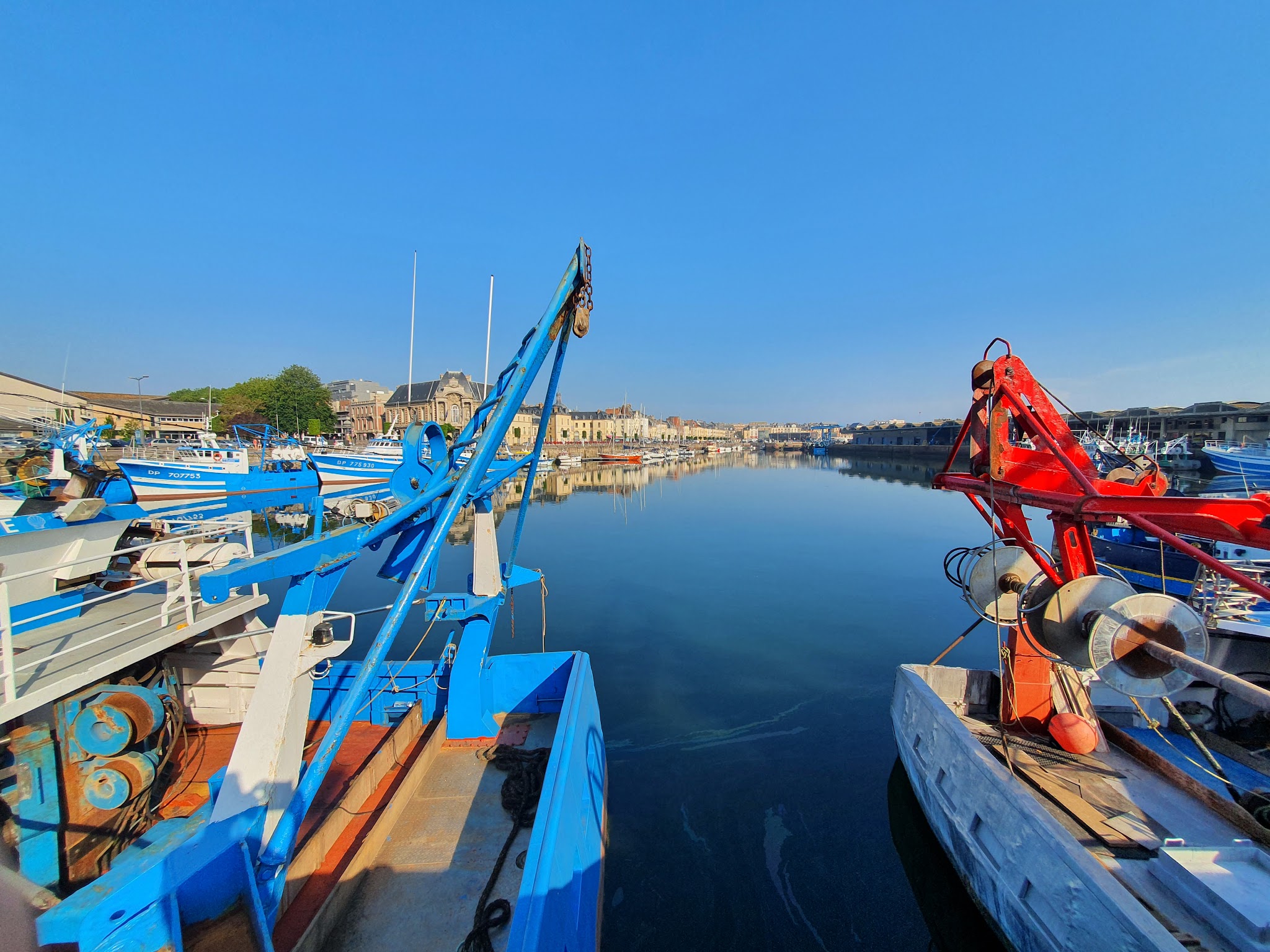 Ruhiger Hafen unter blauem Himmel; blau-weiß gestreifte Fischerboote spiegeln sich im klaren Wasser.