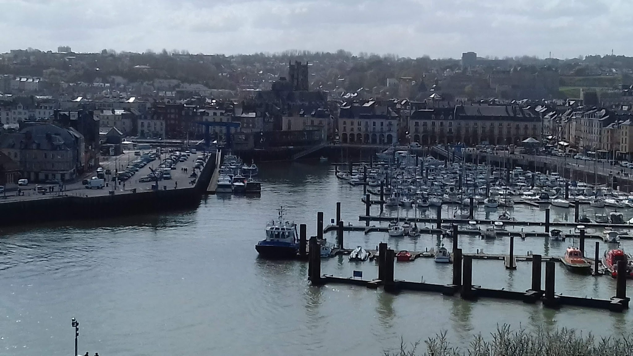 Städtischer Hafen voller Segelboote, Pier mit Autos, historische Häuser und Kirche im Hintergrund.