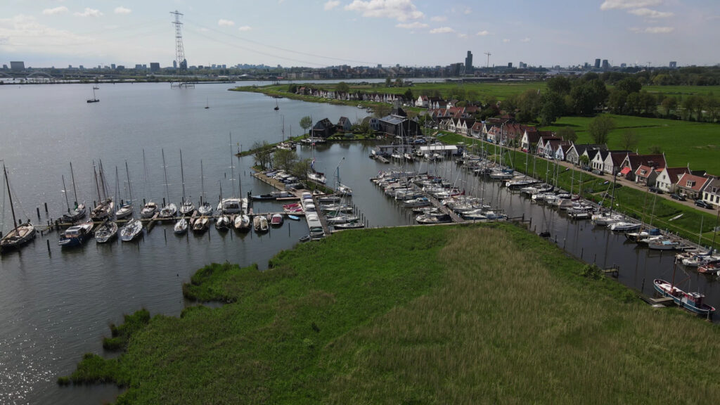 Luftaufnahme eines Yachthafens voller Segelboote, bunte Giebelhäuser entlang des Kanals, grüne Wiesen, ferne Skyline