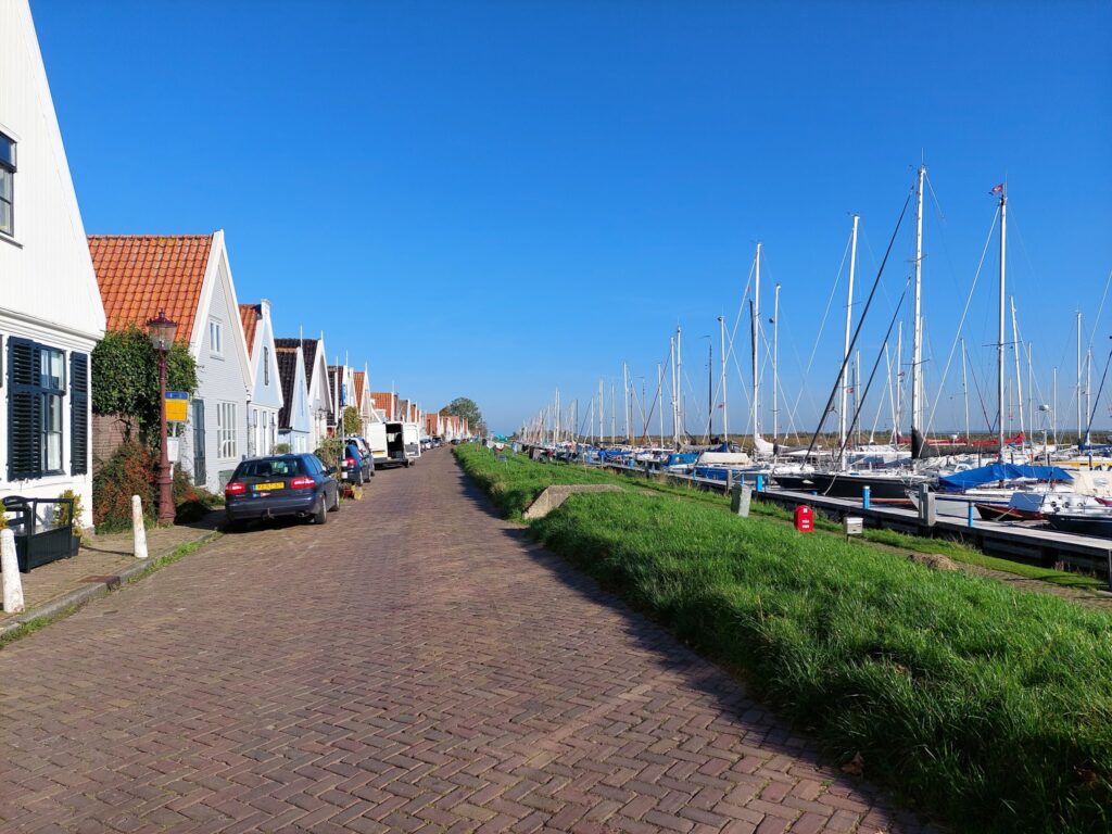 Kopfsteinpflastergasse mit Giebelhäusern links, Yachthafen voller Segelboote rechts, alles unter wolkenlosem Himmel