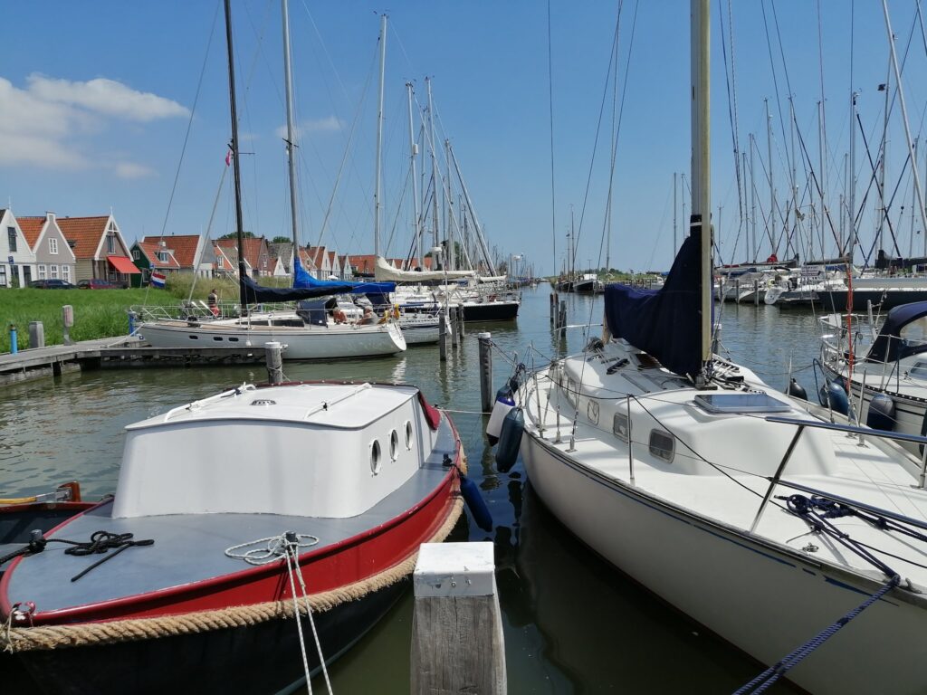 Sonniger Yachthafen mit mehreren Segelbooten, rotbedachten Giebelhäusern im Hintergrund und blauem Himmel.