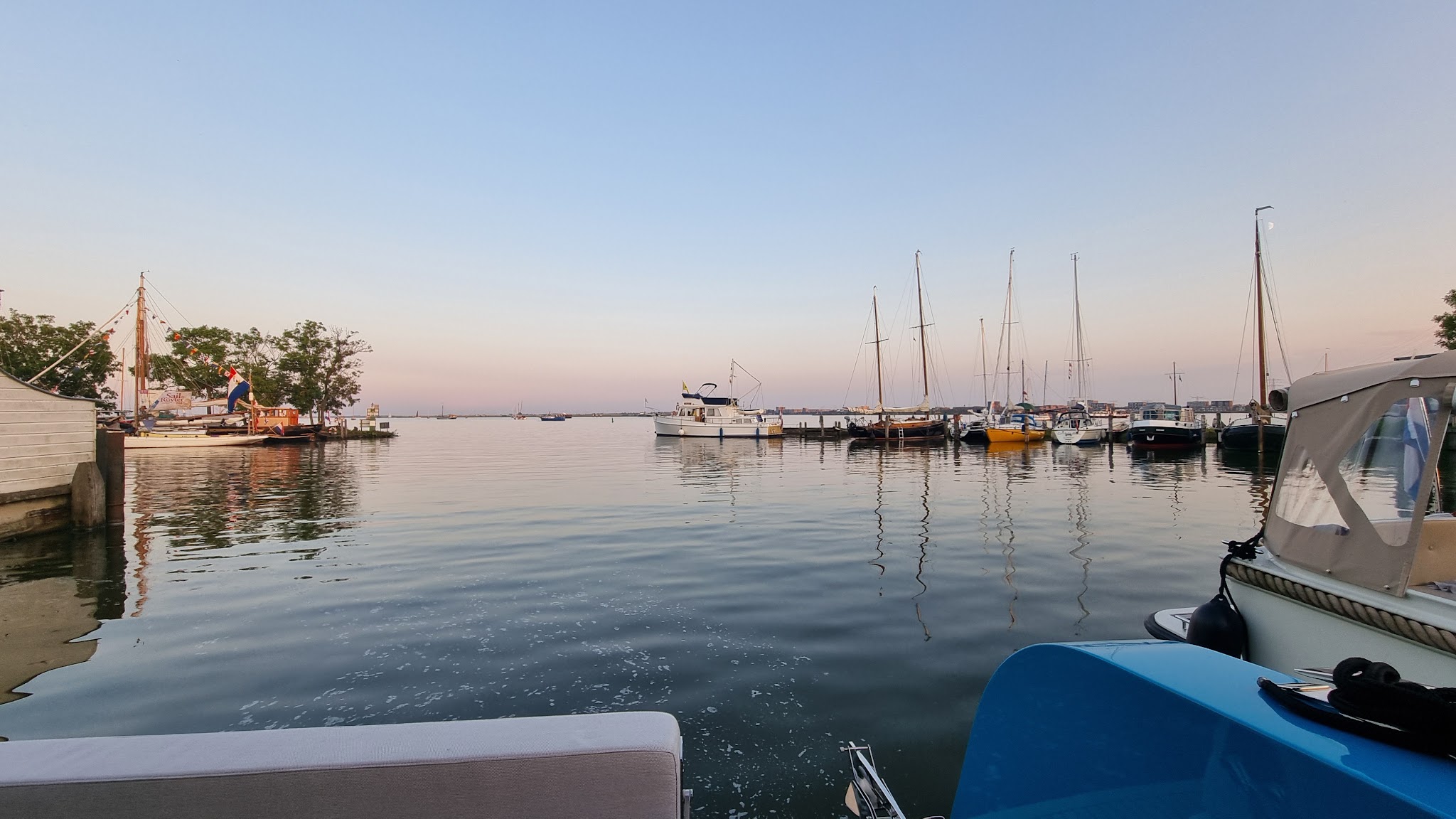 Ruhiger Hafen bei Sonnenuntergang: Segel- und Motorboote an Stegen, glattes Wasser spiegelt den Pastellhimmel.