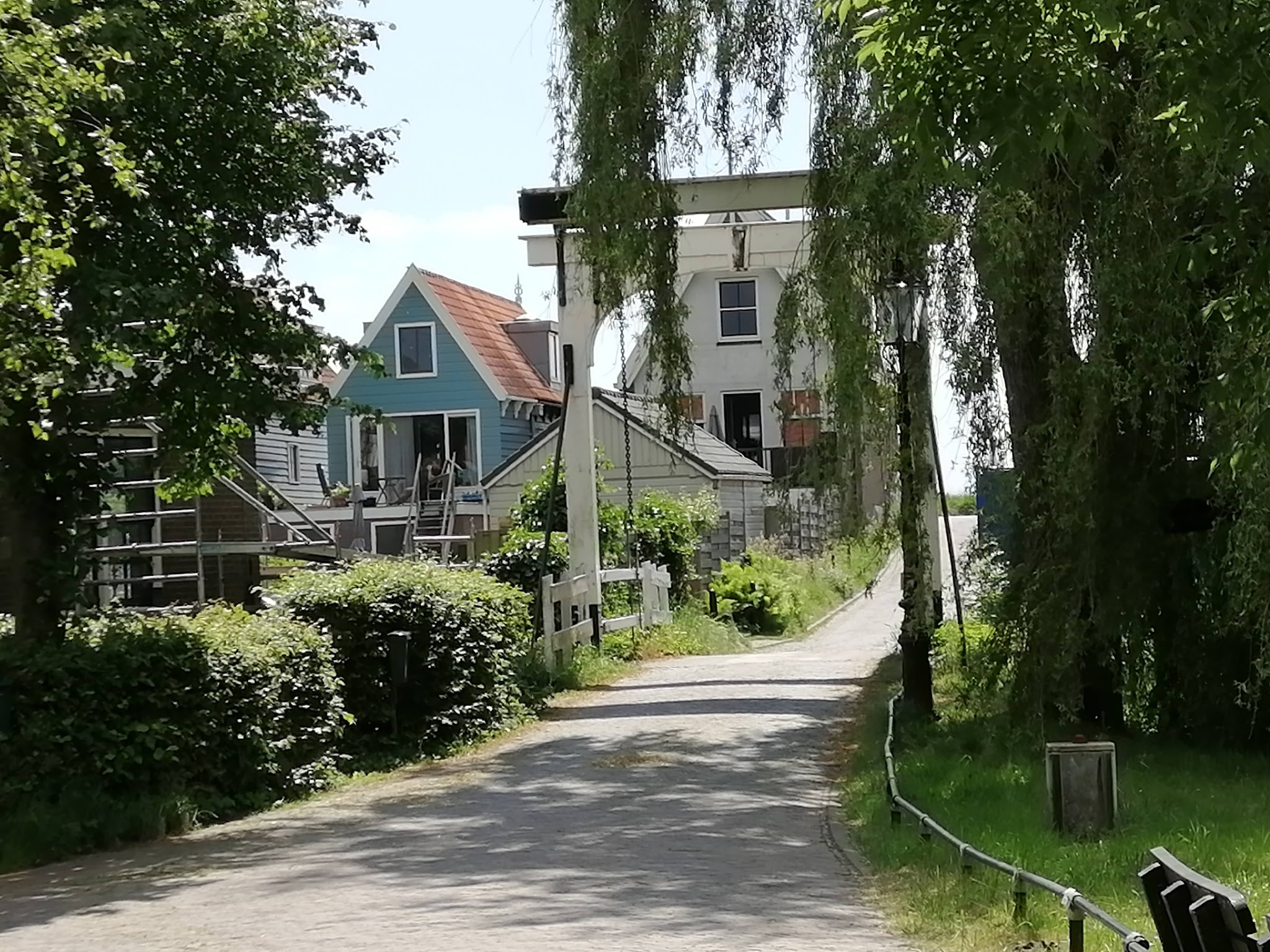 Baumgesäumter Weg führt über weiße Klappbrücke zu bunten Häuschen am Kanal, Sommerlicht und Grün.