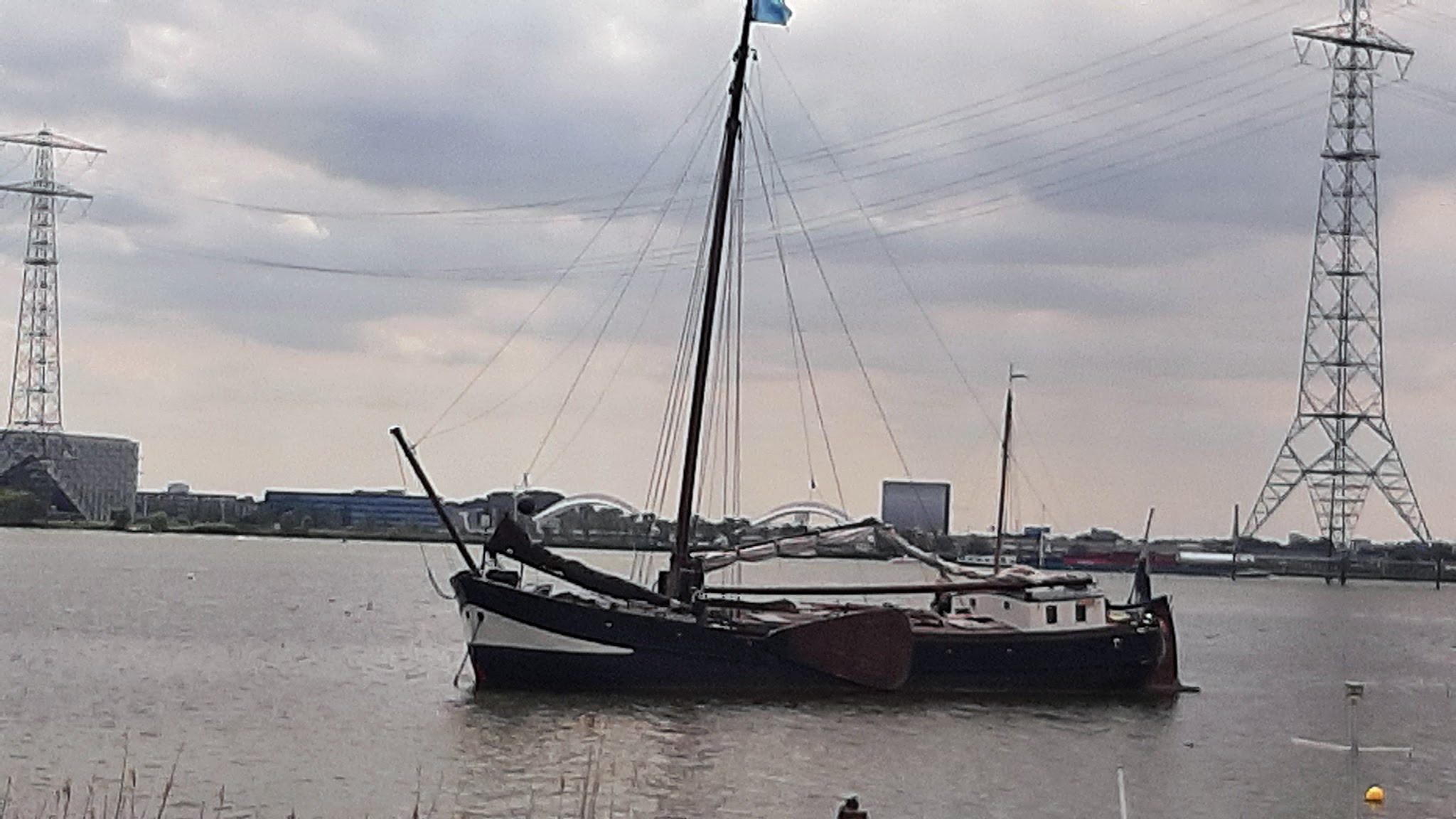 Traditionelles Segelboot liegt auf ruhigem Fluss; im Hintergrund Brückenbogen, Industrie und Strommasten unter Wolken.
