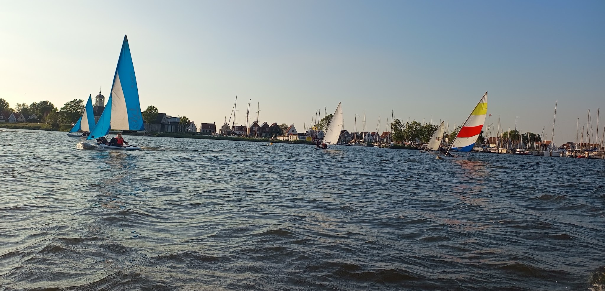 Mehrere Segelboote mit bunten und blauen Segeln kreuzen am Abend vor einem Hafen mit Häusern und Masten.