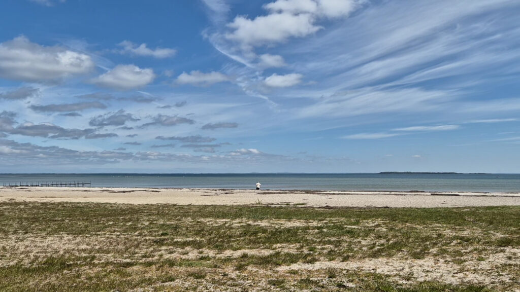 Weite Ostseestrand­landschaft unter blauem Himmel; einzelne Person am Wasser, Steg links, Insel am Horizont.