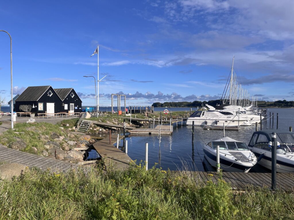 Kleiner Yachthafen mit weißen Segel- und Motorbooten, schwarzem Bootshaus, Holzsteg und klarem blauen Himmel.