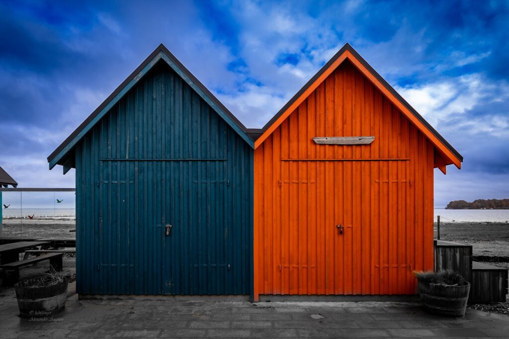 Zwei Holzstrandhütten nebeneinander: links tiefblau, rechts leuchtend orange, vor wolkigem Himmel und Meer.