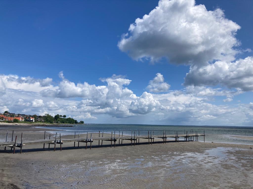 Holzsteg führt über wattähnlichen Sandstrand ins ruhige Meer; dahinter Küstenhäuser, weite Wolken am blauen Himmel