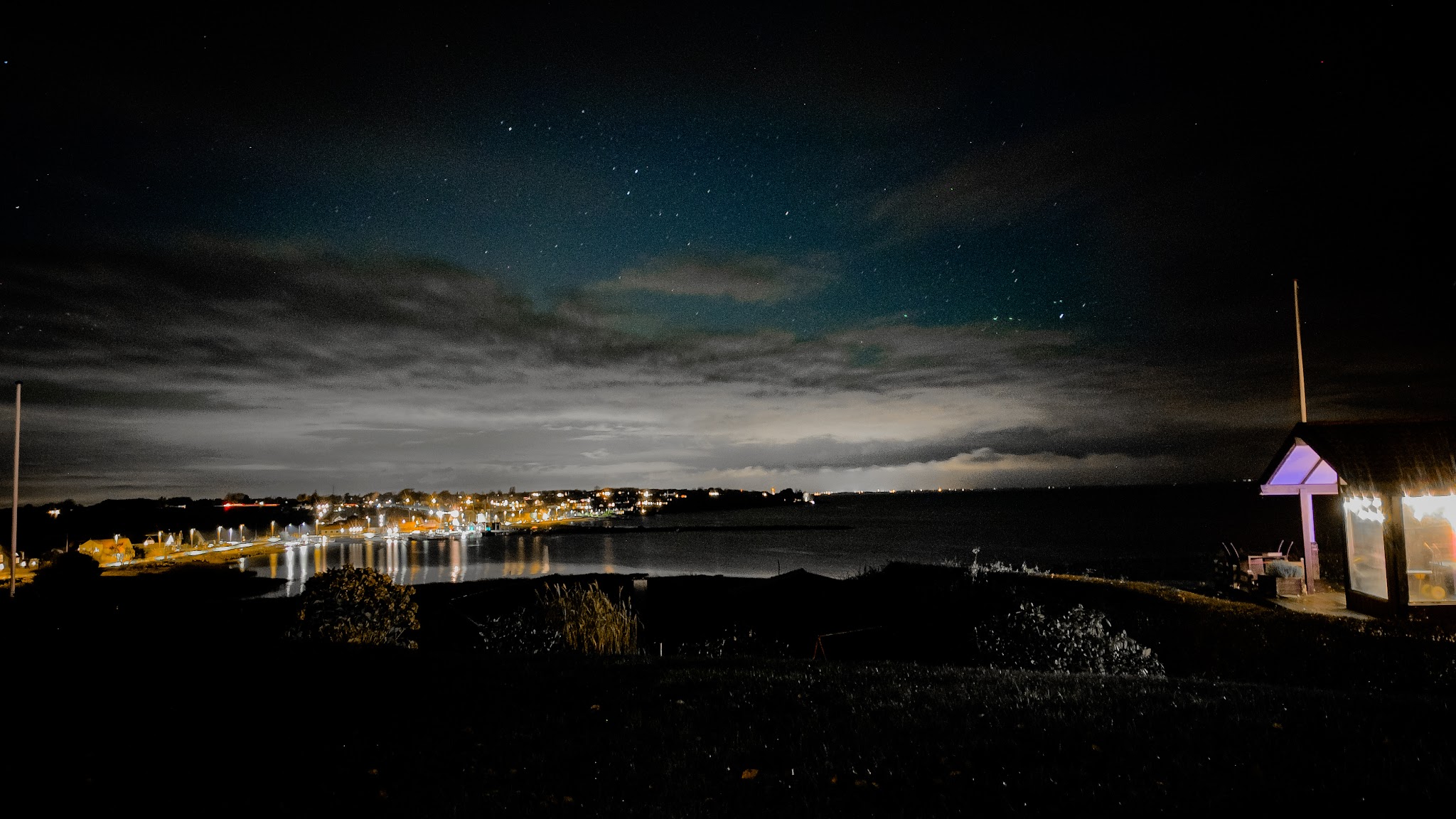 Nachtküste mit Stadtlichtern an ruhiger Bucht, Sternenhimmel, rechts ein lila beleuchtetes Strandhäuschen.