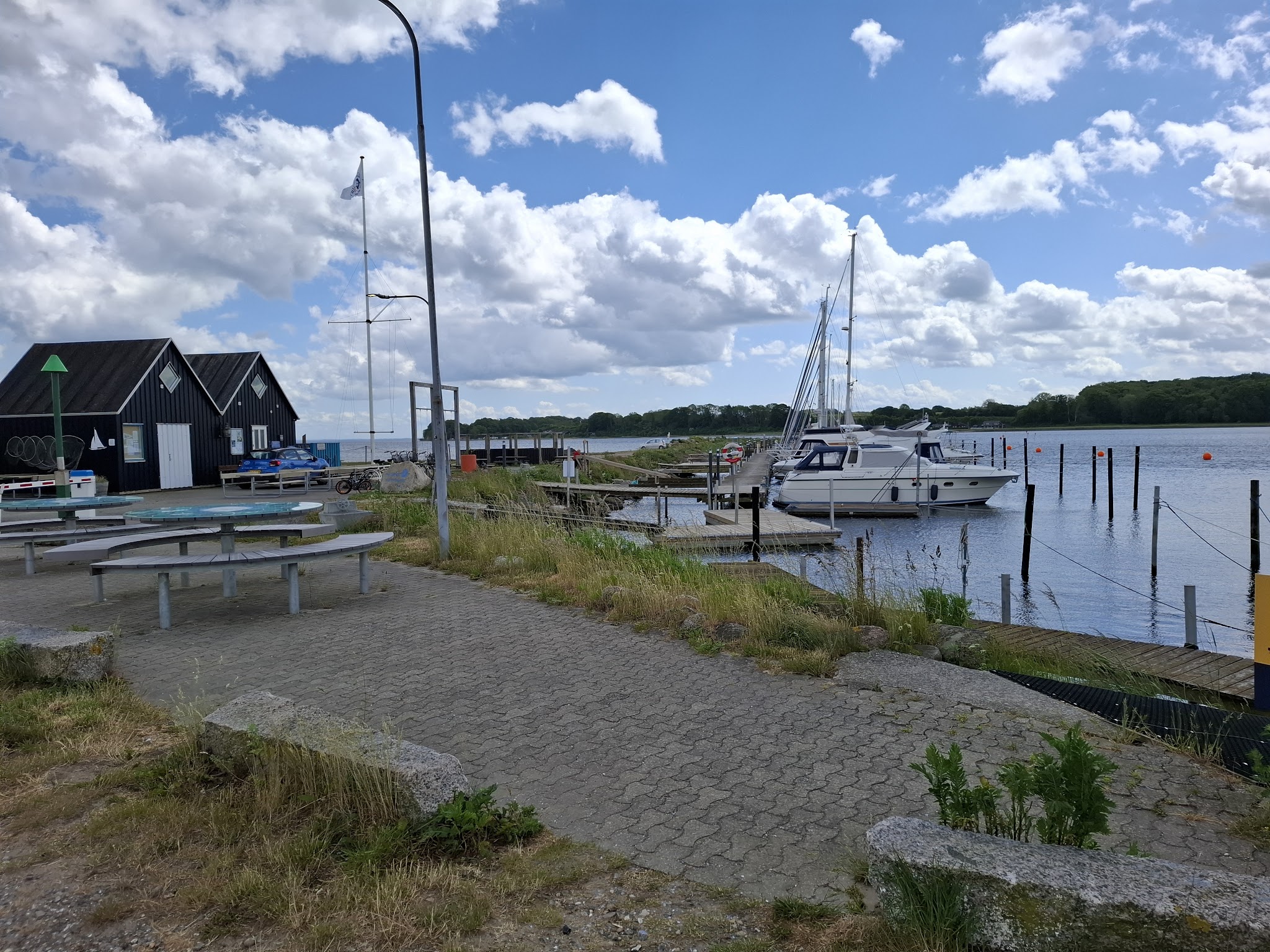 Kleiner Jachthafen mit festgemachter Motoryacht, schwarzen Bootshäusern und runden Picknicktischen unter Wolkenhimmel