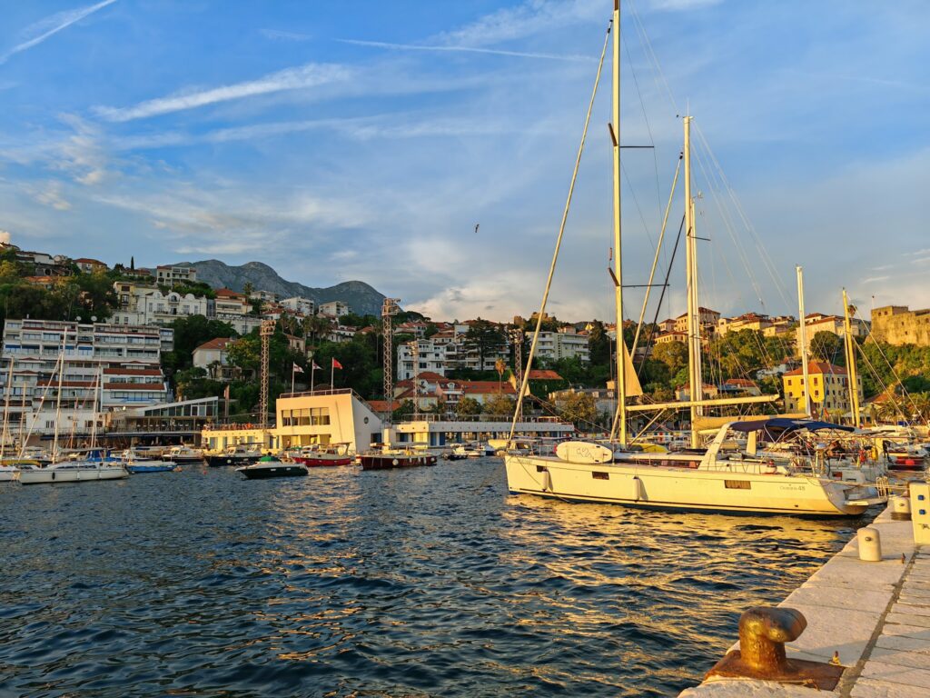 Segelboote im Yachthafen vor hügeliger Küstenstadt, Abendsonne färbt Wasser und Häuser golden