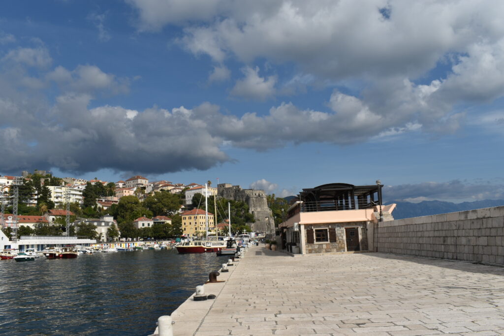 Hafenpromenade mit Segelbooten; dahinter bunte Hängehäuser und mittelalterliche Festung unter teils wolkigem Himmel.