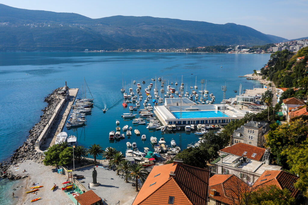 Marina mit vielen Segel- und Motorbooten, langer Pier, großes Meerwasserbecken, rote Dächer, bewaldete Berge im Hintergrund