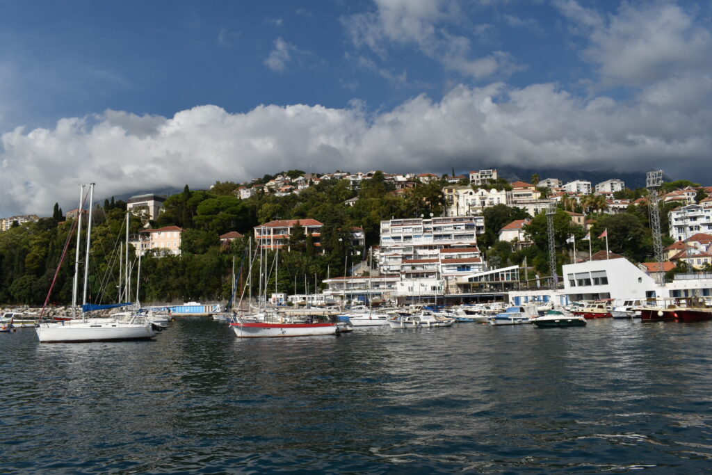 Hafen mit Segelbooten vor grünem Hang voller Häuser; darüber ziehen tiefe Wolken vor blauem Himmel.