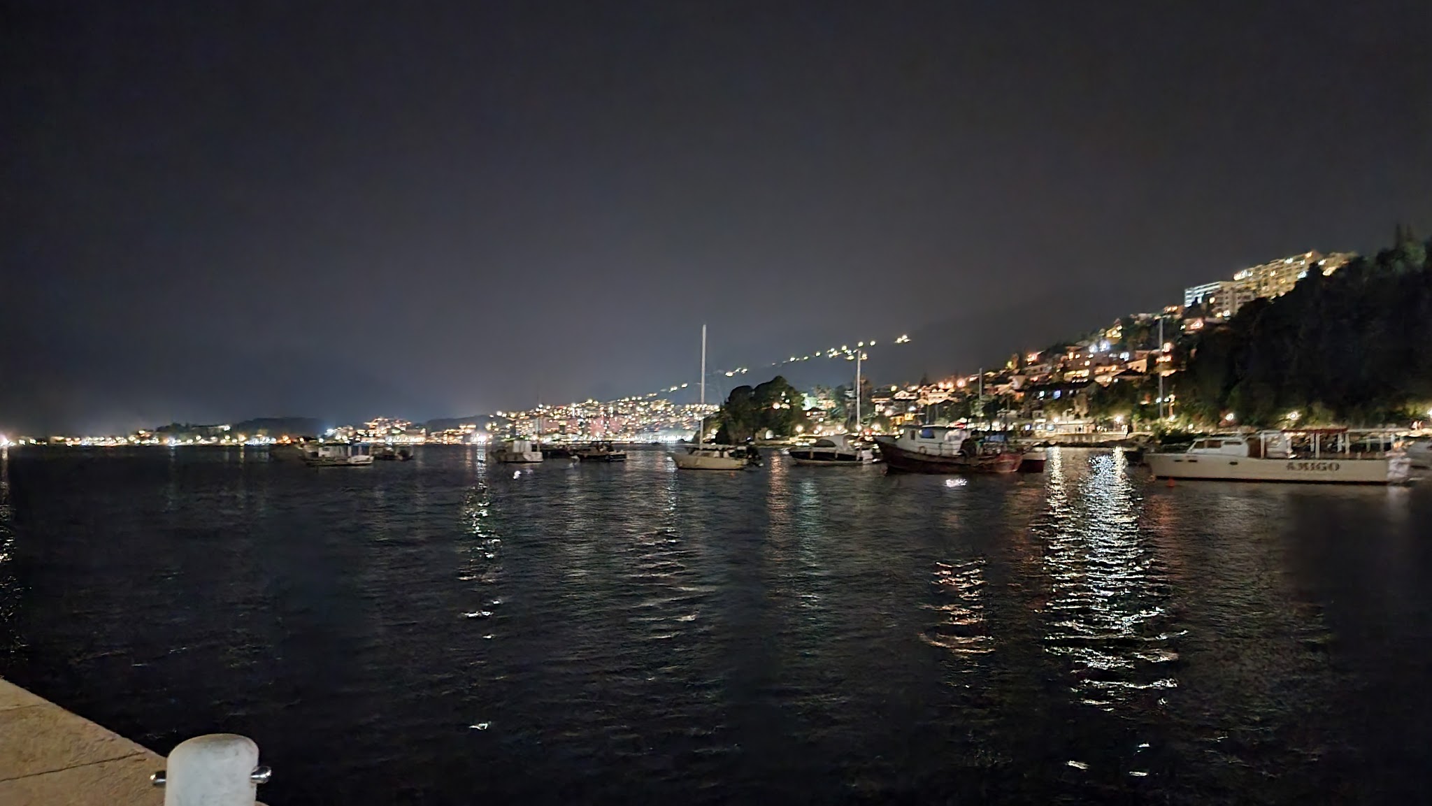 Beleuchteter Küstenhafen bei Nacht, Boote liegen im dunklen Wasser, Stadthang voller Lichter spiegelt sich.