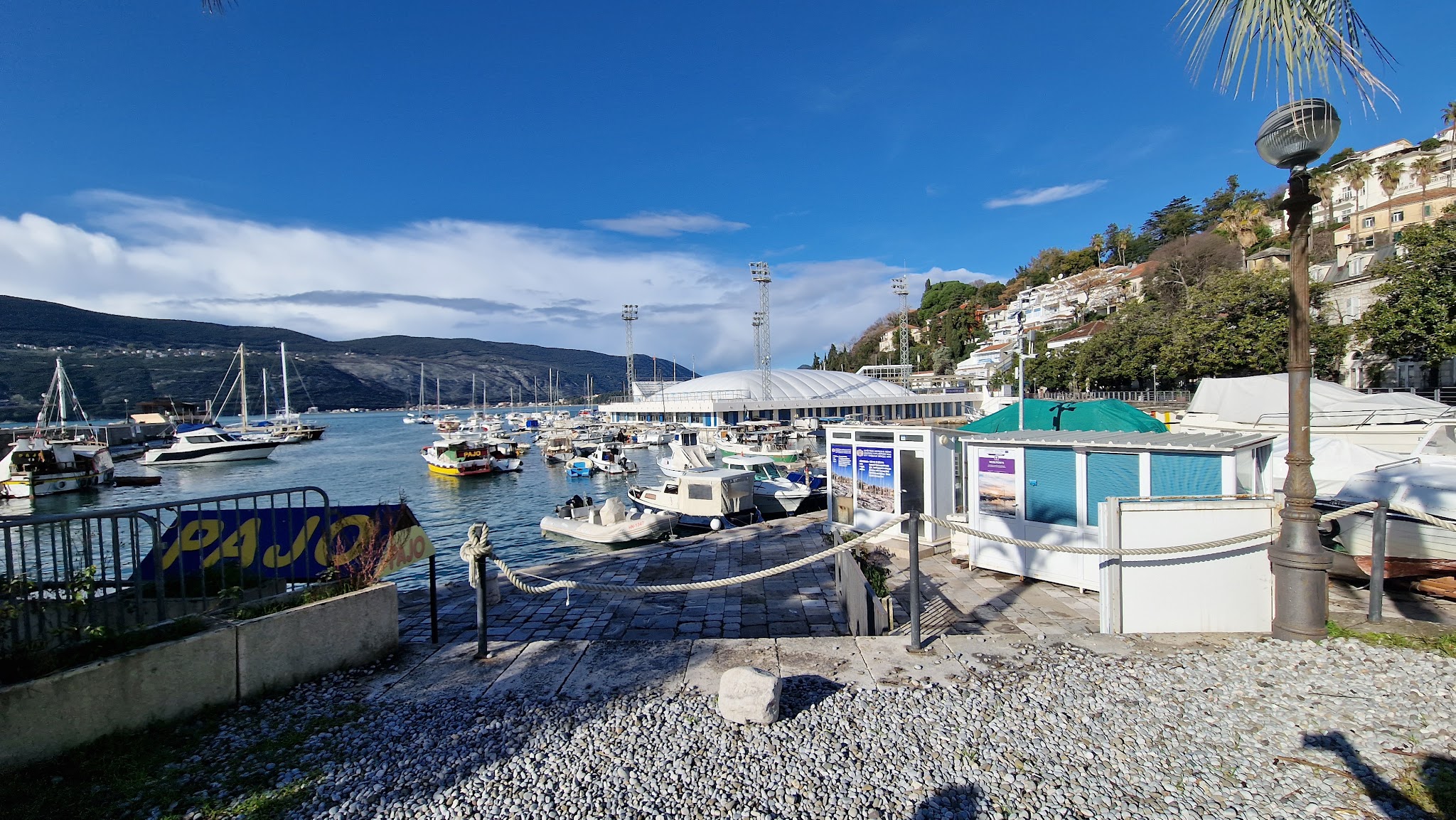 Sonniger Küstenhafen; viele kleine Boote liegen im türkisblauen Wasser vor bewaldeten Hügeln und Häusern.