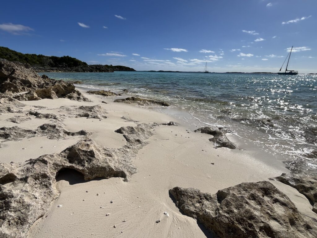Sonniger Strand mit hellem Sand und Felsen, türkisfarbenem Meer und zwei Segelbooten am Horizont