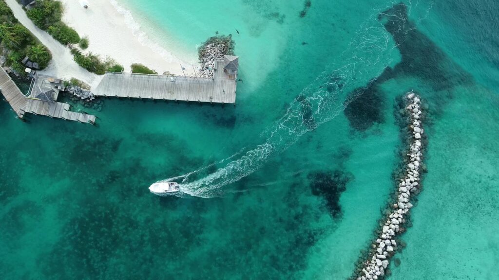Motorboot zieht weiße Spur vom Holzsteg durchs türkis klare Meer; linker Sandstrand, rechts gebogene Steinmole.