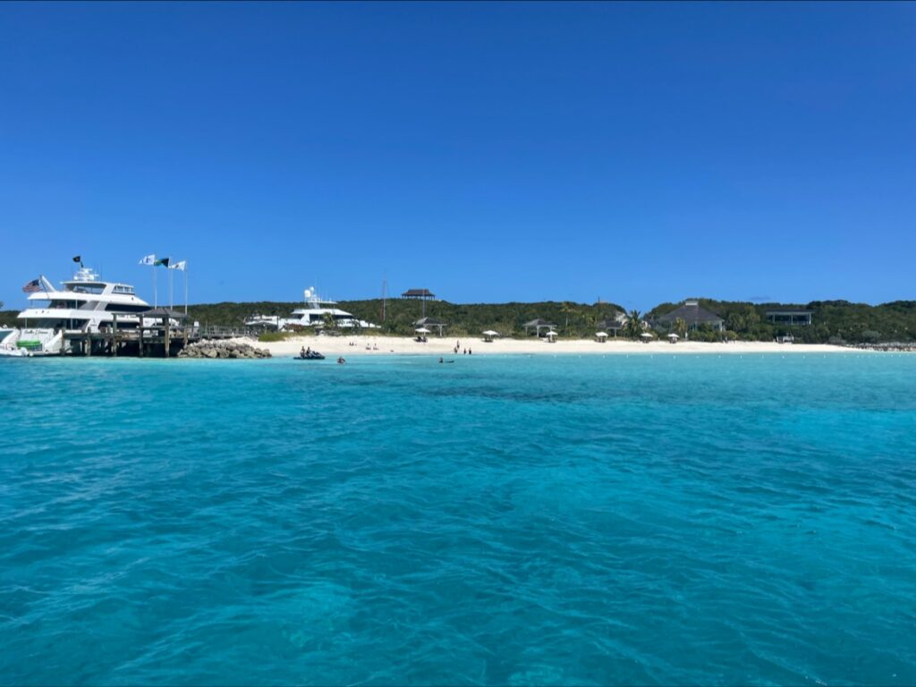 Türkisblaues Meer vor weißem Sandstrand; Yachten am Steg, wenige Badegäste, grüner Hügel unter klarem Himmel.