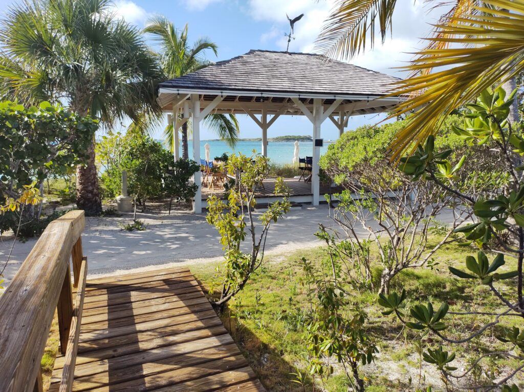 Holzsteg führt durch Palmen zu einem offenen Pavillon am türkisblauen Meer