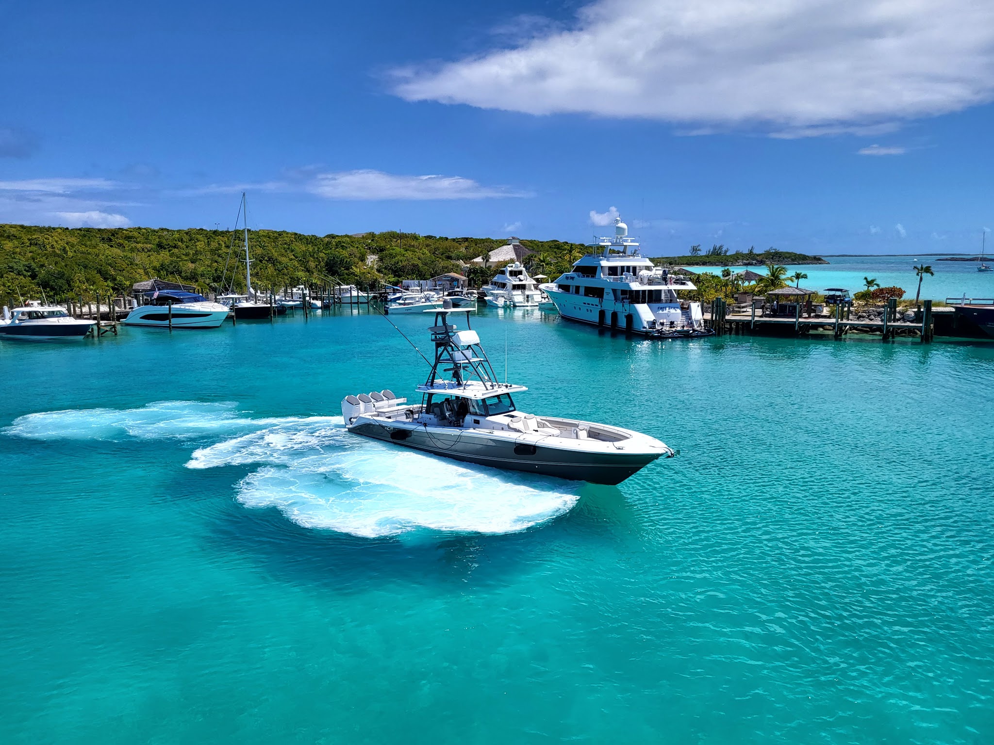Schnelles Motorboot zieht weiße Gischt im türkisfarbenen Hafen; mehrere Yachten liegen vor grün bewachsenen Inseln.