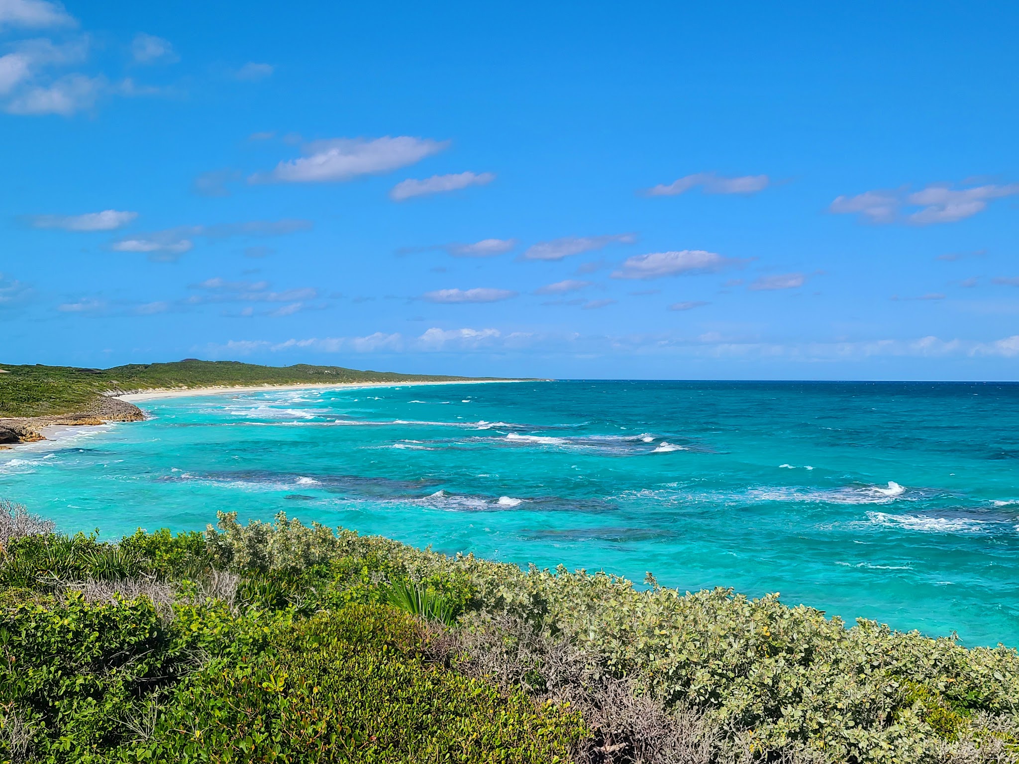 Türkisblaue Küste mit sanften Wellen, langem Sandstrand und dichtem grünen Buschwerk unter klarem Himmel.