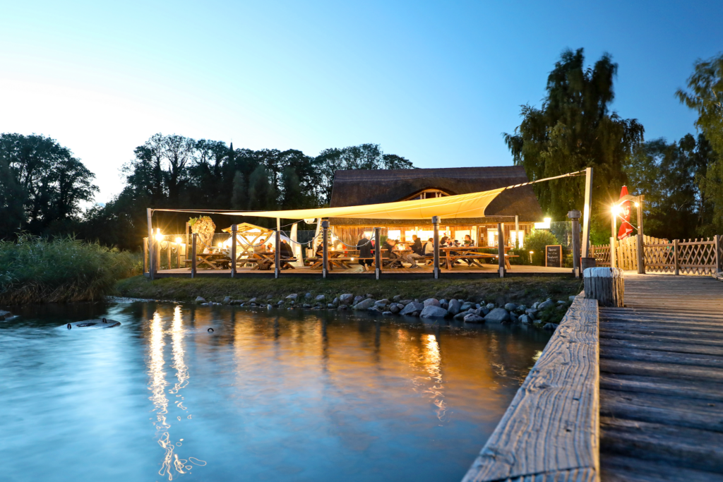 Abendstimmung: beleuchtetes Seerestaurant mit Sonnensegel, Gästen an Holzbänken, Lichtspiegelung im Wasser, Steg rechts.
