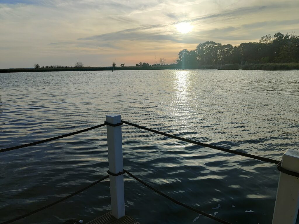 Sonnenuntergang über ruhigem See; Wasser spiegelt Licht, Steg mit weißen Pfosten und Seil im Vordergrund.