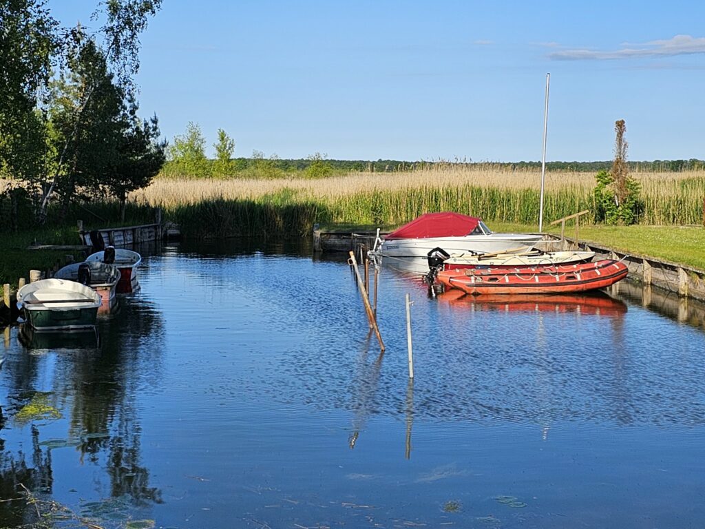 Kleiner Kanal mit mehreren festgemachten Ruder- und Motorbooten, umgeben von Schilf und Bäumen unter blauem Himmel.