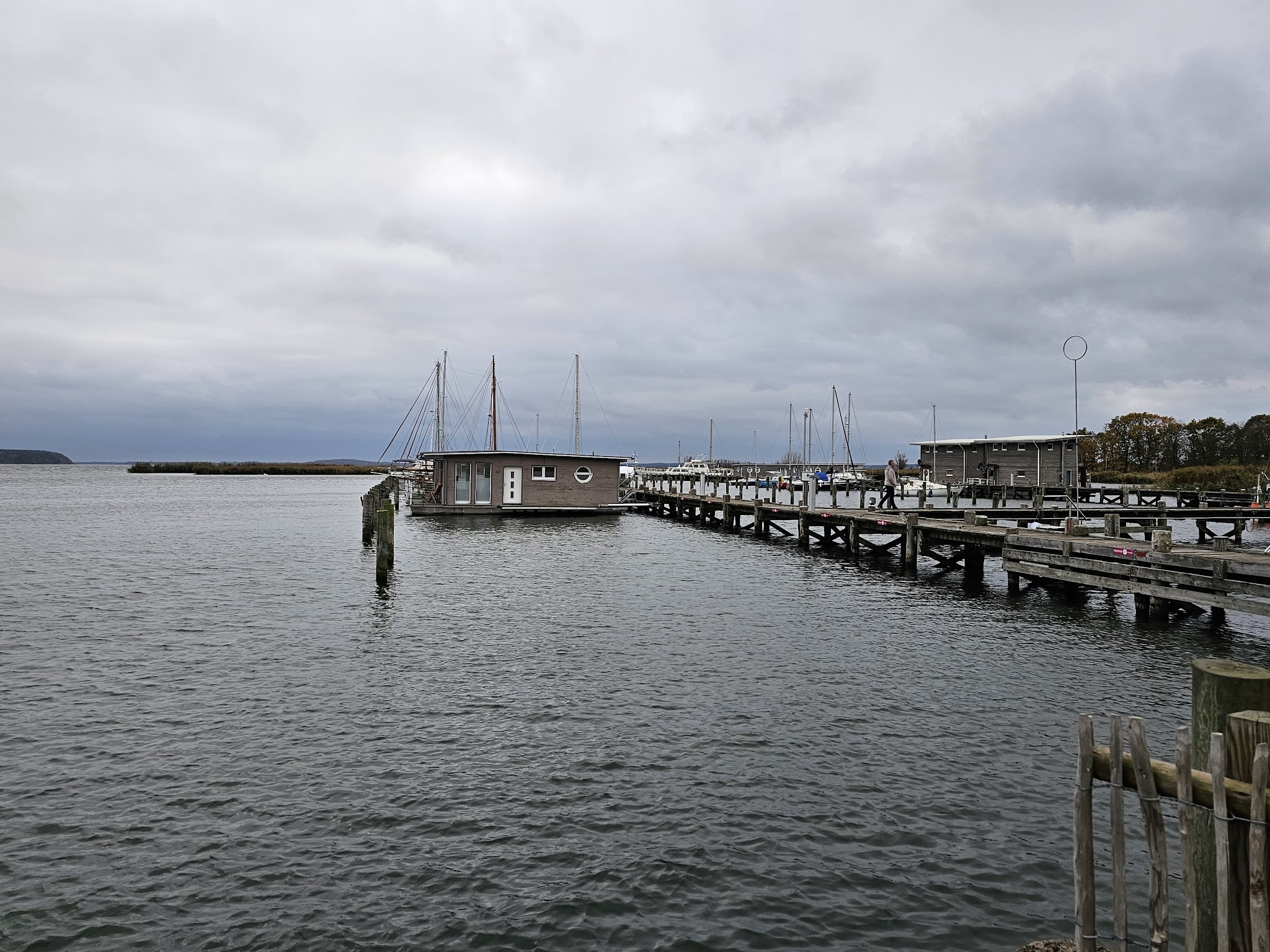 Langer Holzsteg mit Hausboot und Segelmasten ragt in ruhige Bucht unter bewölktem Himmel.
