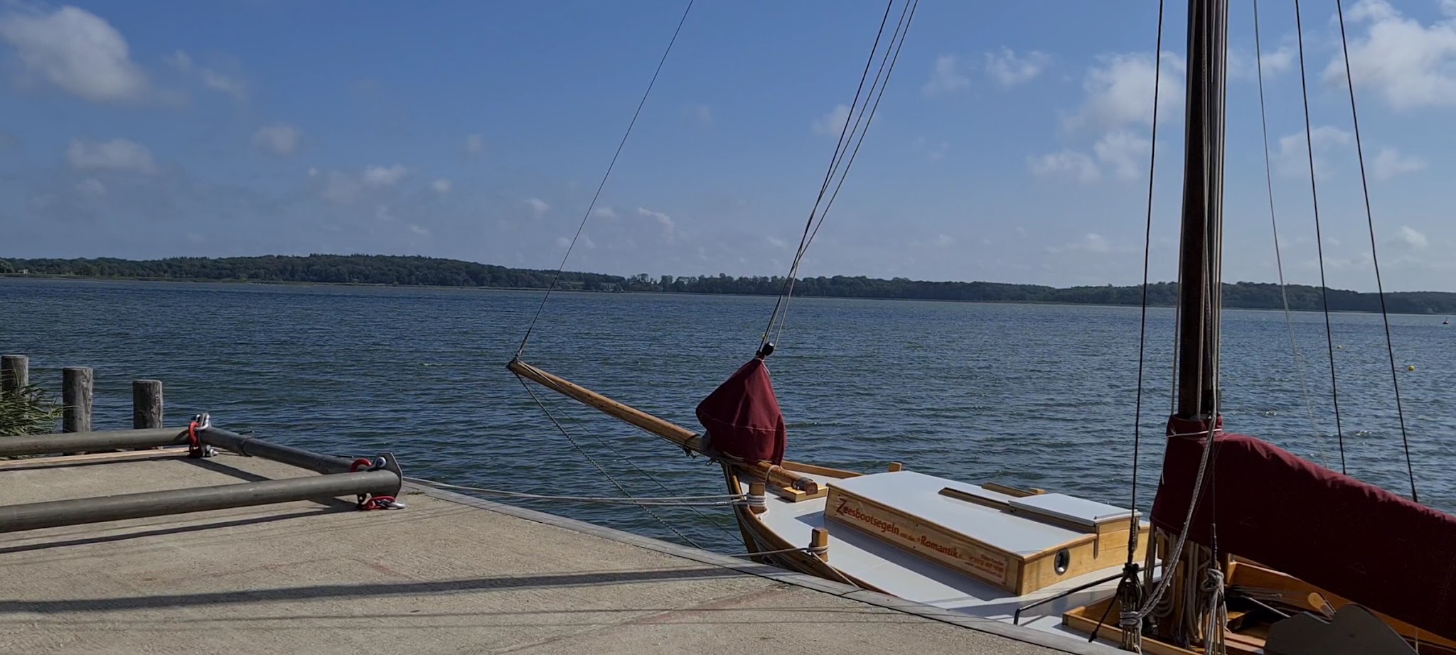 Holzsegelboot mit eingezogenem Mast vertäut am Steg, ruhiger See und bewaldetes Ufer unter blauem Himmel.