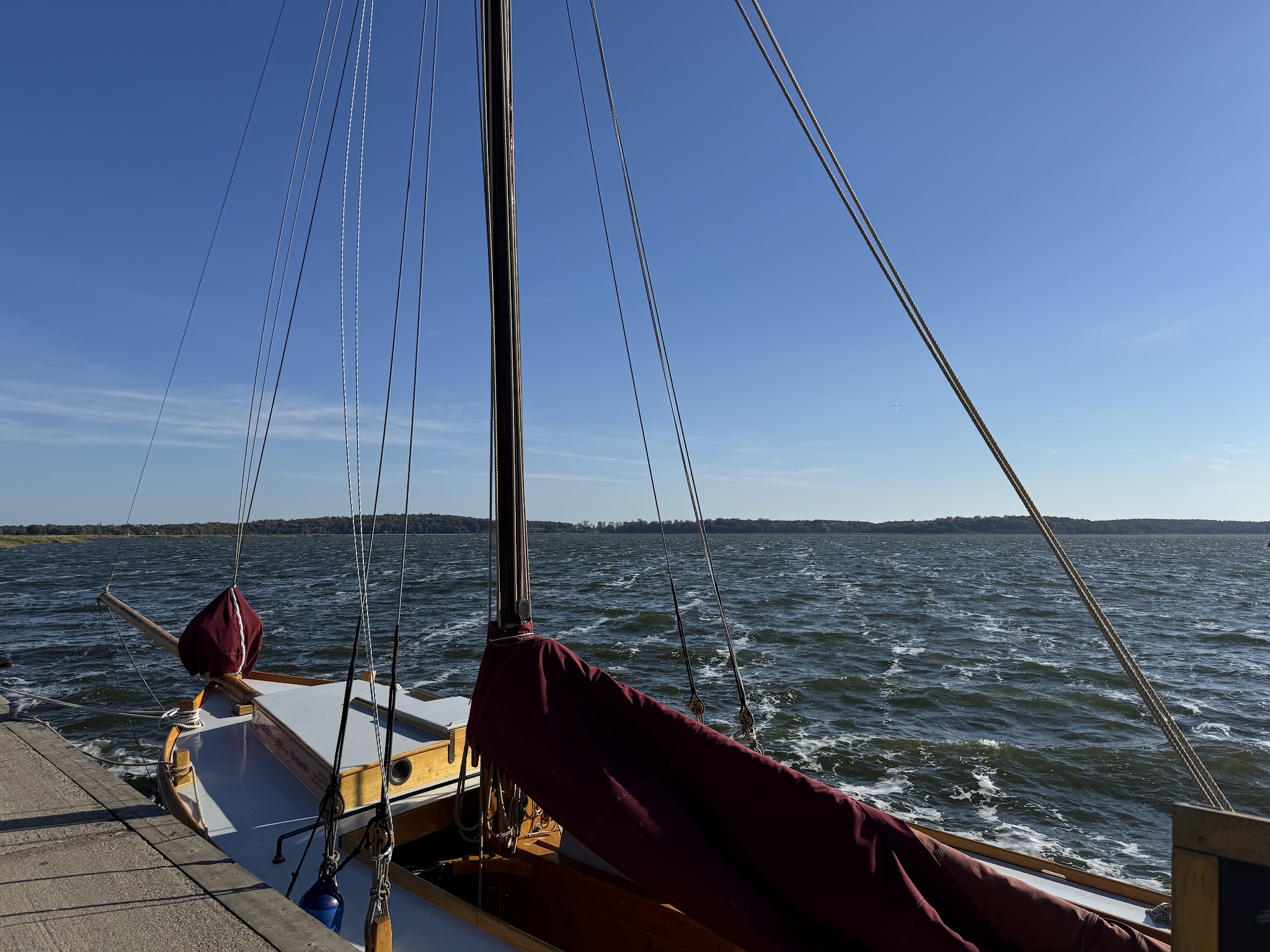 Vertäutes Segelboot am Steg, hohe Masten und Leinen vor windgekräuseltem See unter klarem blauem Himmel