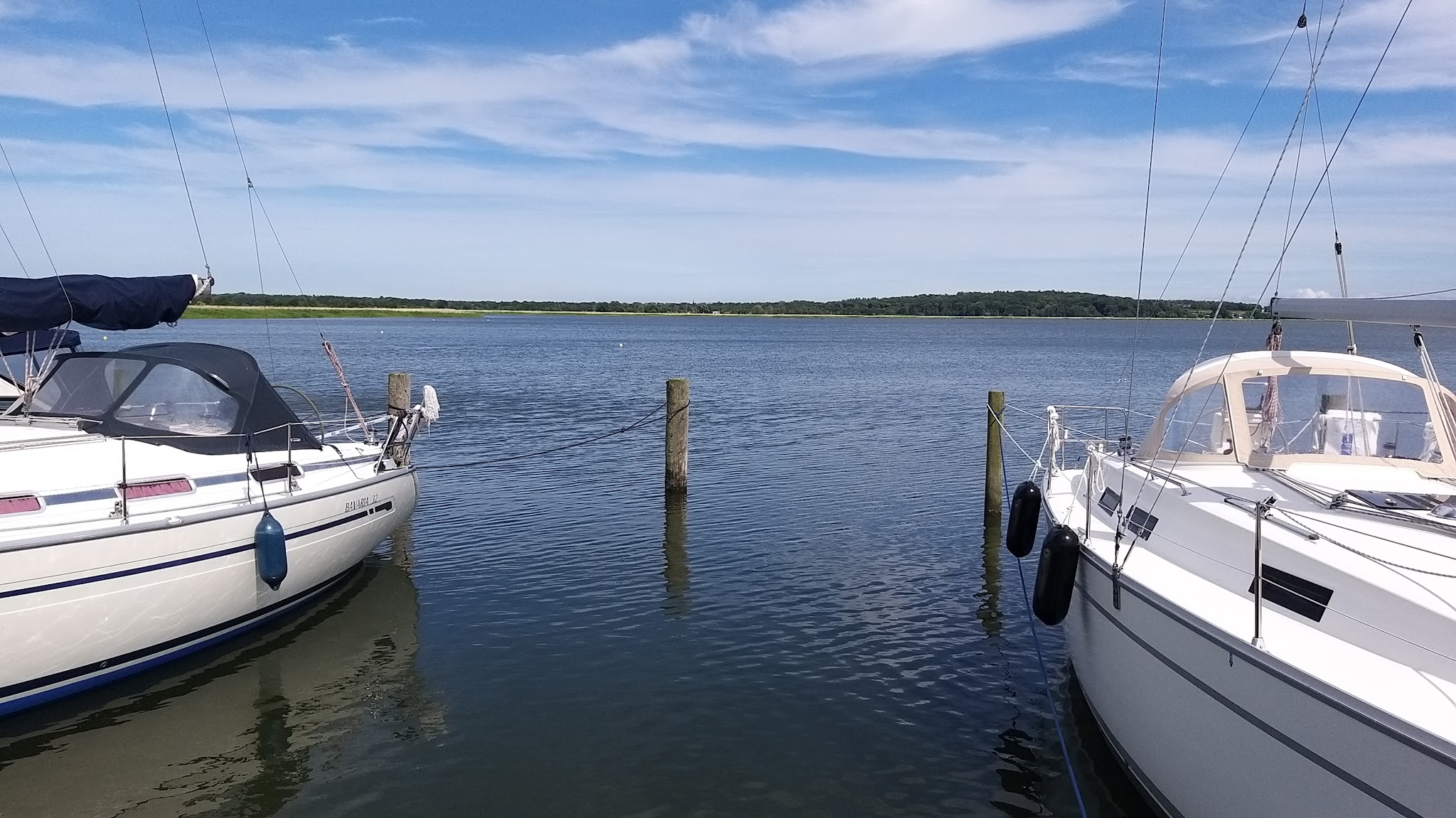 Zwei Segelboote liegen an Pfählen auf ruhigem See; blauer Himmel und bewaldetes Ufer im Hintergrund.