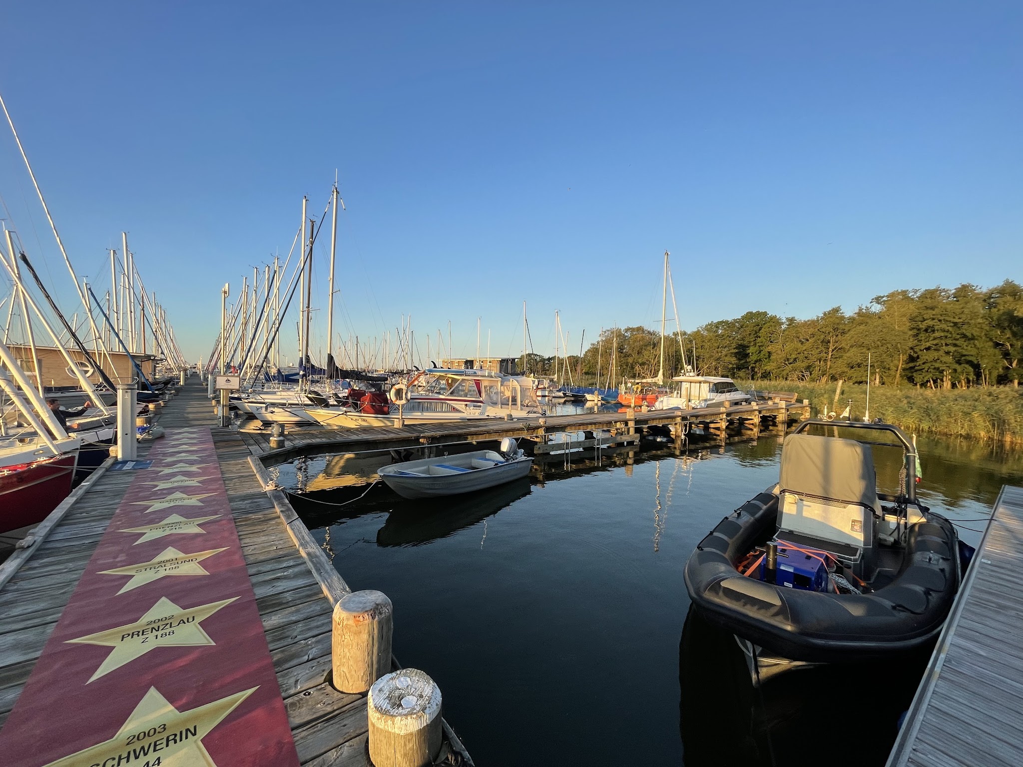Steg mit rotem Teppich voller Stern-Motiven, links und vorn viele Segelboote, ruhiges Hafenbecken im Abendlicht.