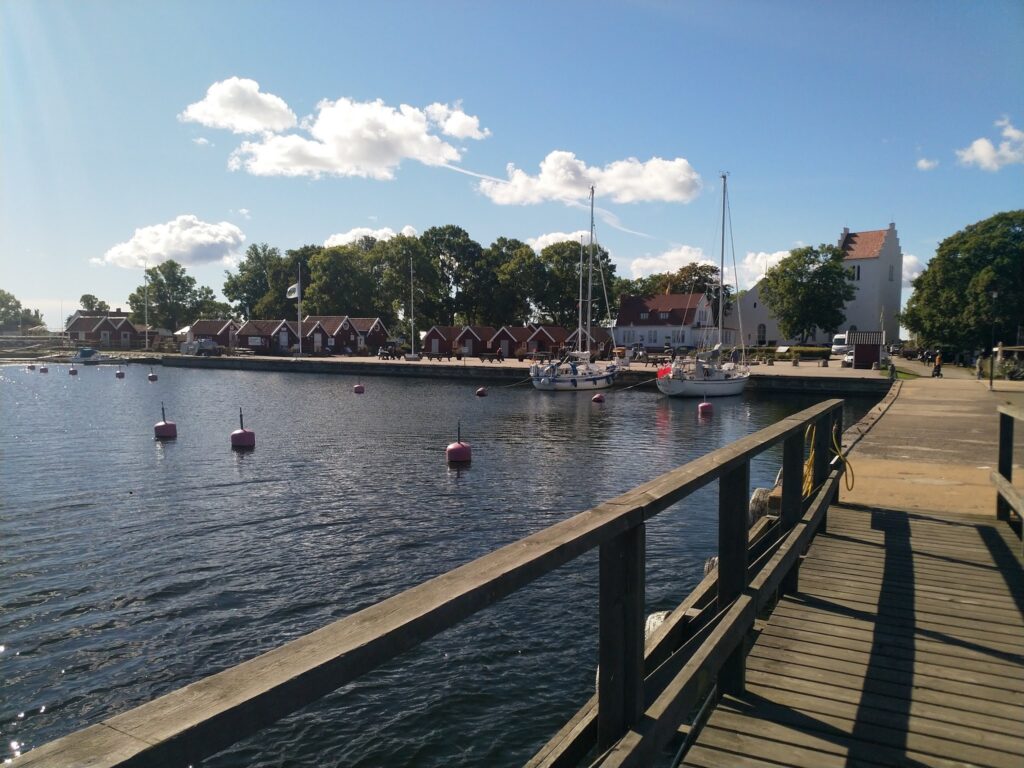 Sonniger kleiner Hafen mit roten Fischerhütten, Segelbooten, weißer Kirche und Holzsteg über ruhigem Wasser.