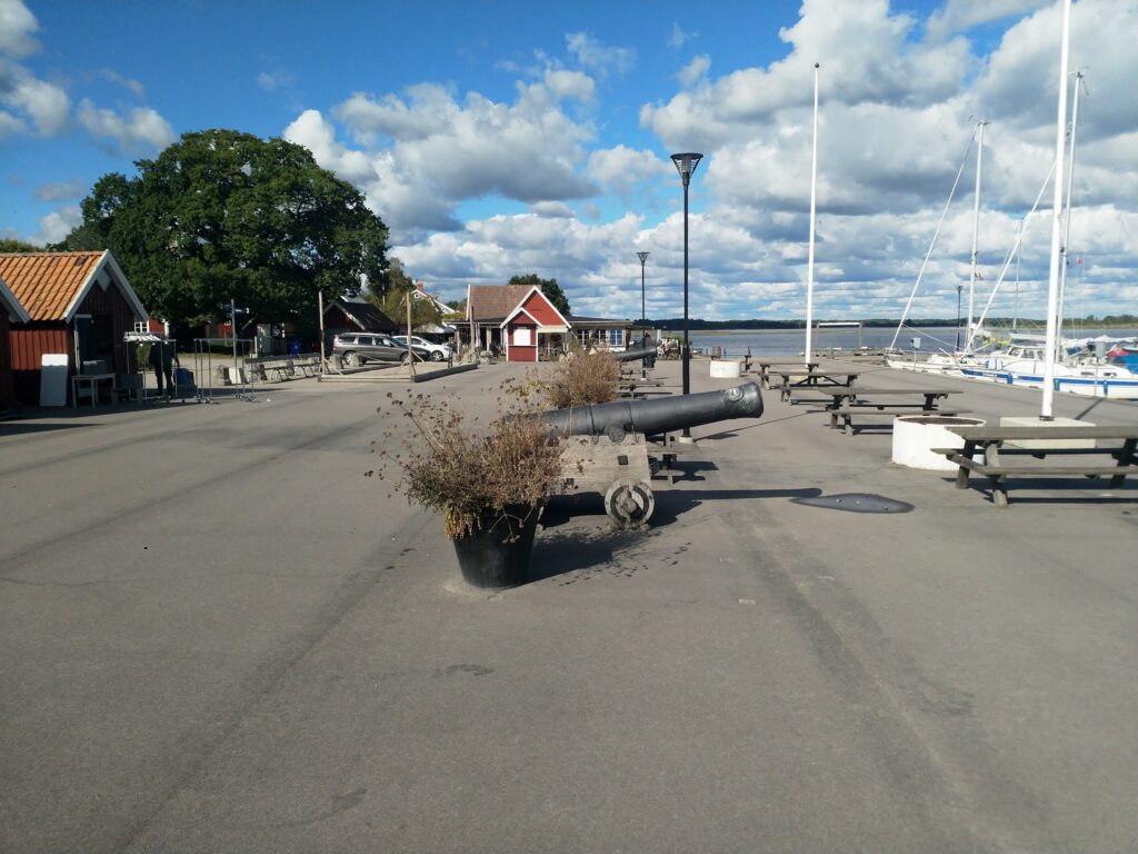Historische Kanone auf Hafenpier mit Segelbooten, roten Holzhütten und blauem, wolkigem Himmel.