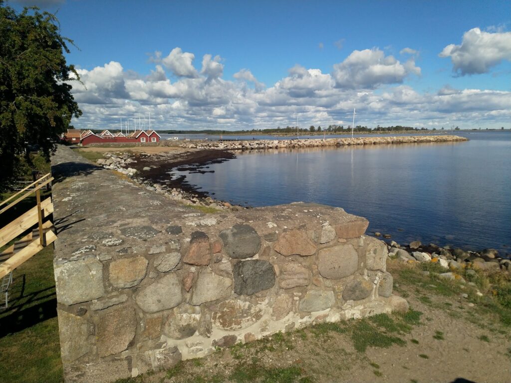 Steinmauer am ruhigen Küstenwasser, gegenüber rote Bootshäuser; blauer Himmel mit weißen Wolken