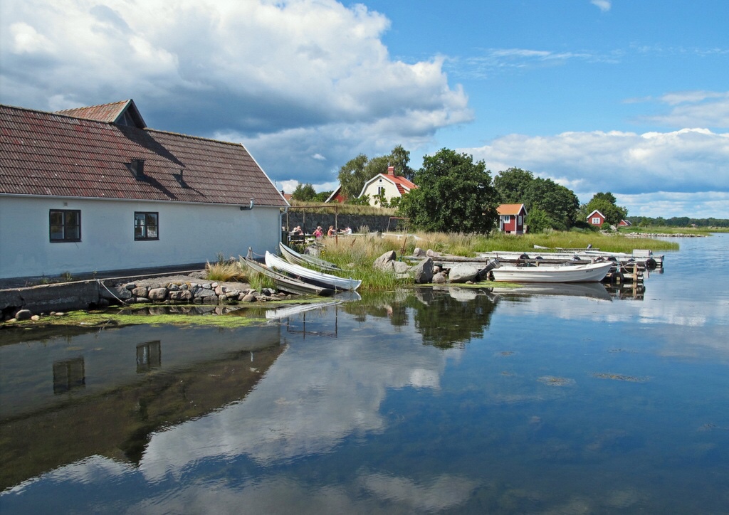 Ruhiger See mit weißem Bootshaus, mehreren Ruderbooten am Steg, rote Häuschen und Wolken spiegeln sich im Wasser.