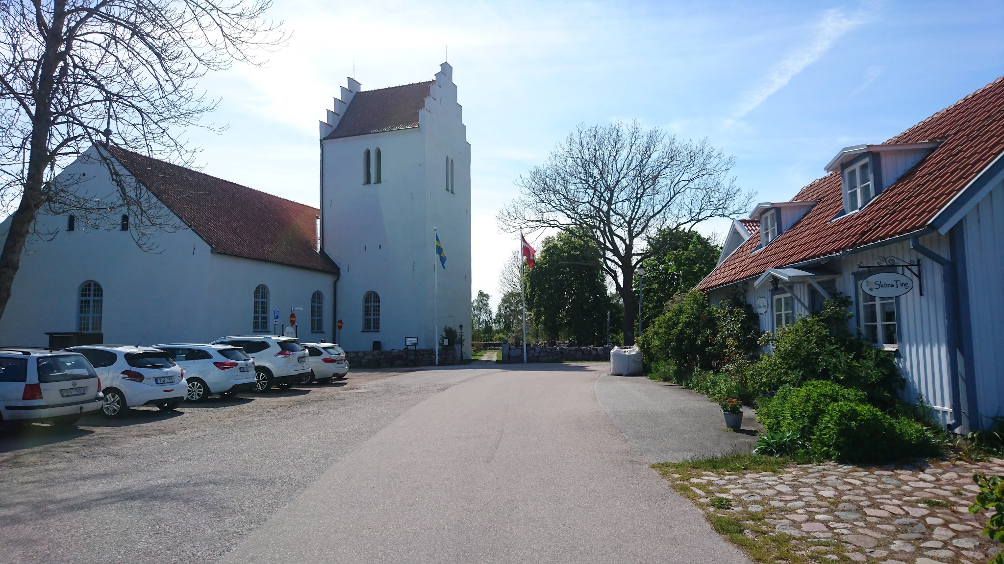 Kleine Dorfstraße mit blauer Kirche, schwedischer Flagge, parkenden Autos und blauem Holzhaus unter klarem Himmel.